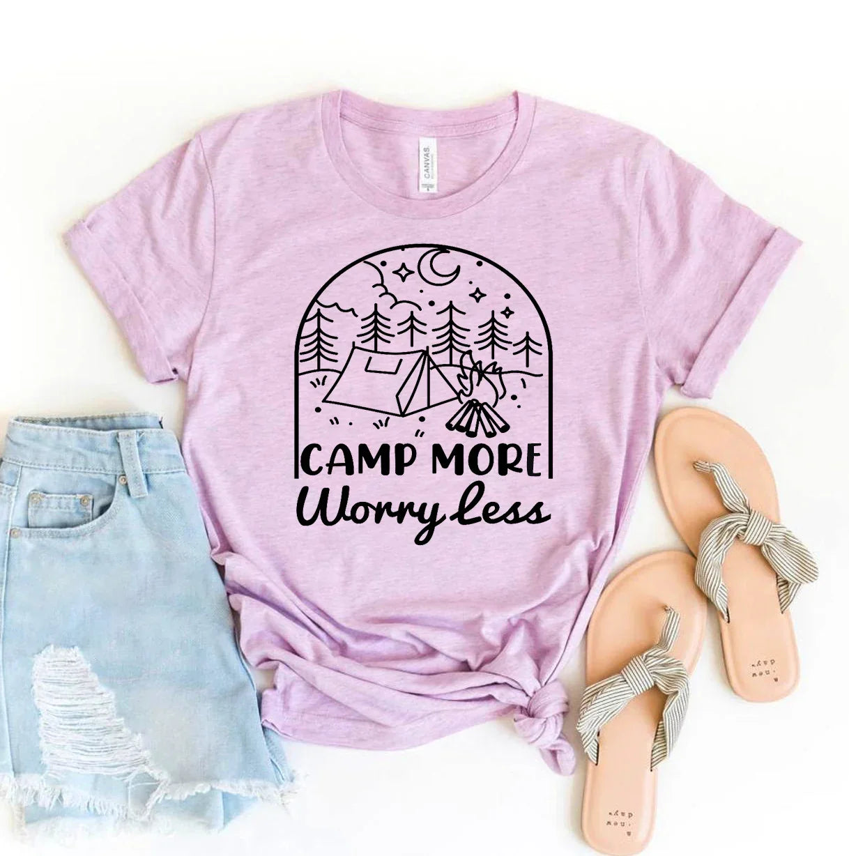 Light purple t-shirt with camping graphic and text 'Camp More Worry Less' laid flat with ripped denim shorts and striped bow sandals