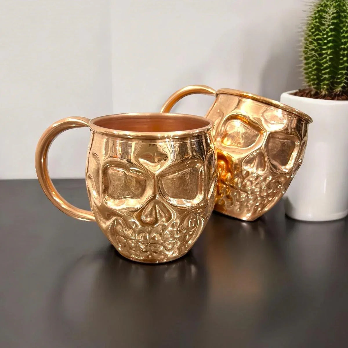 Two metallic skull mugs on a dark surface beside a small potted cactus at Qu!rk Boutique