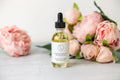 Glass dropper bottle of lavender body oil with pink peony flowers on marble surface