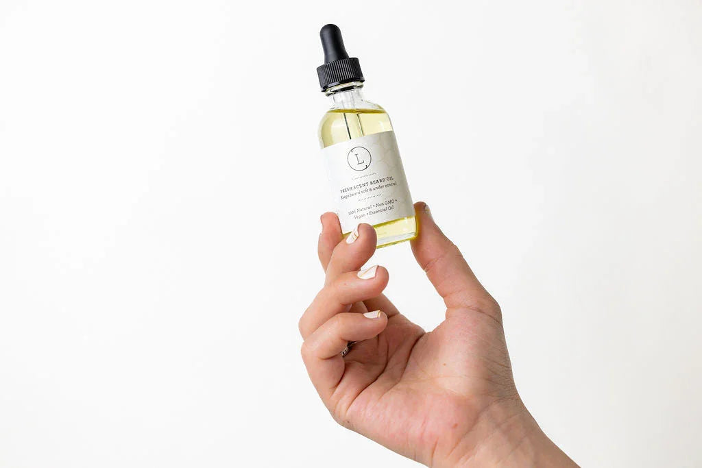 Hand holding a clear glass dropper bottle of natural beard oil with a white label against a white background