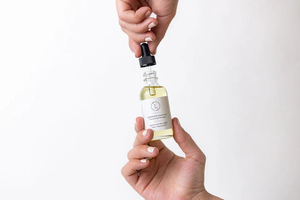 Hands holding a dropper bottle of lavender body oil with a white label on a white background