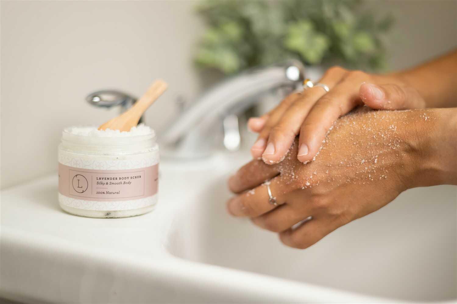 Hands applying lavender body scrub over sink with jar of natural exfoliating scrub