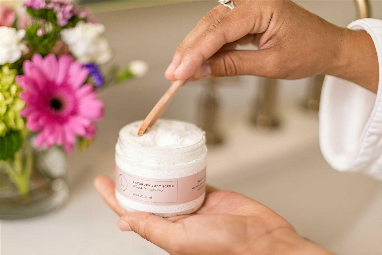 Hands holding and scooping lavender body scrub from jar with flowers in background