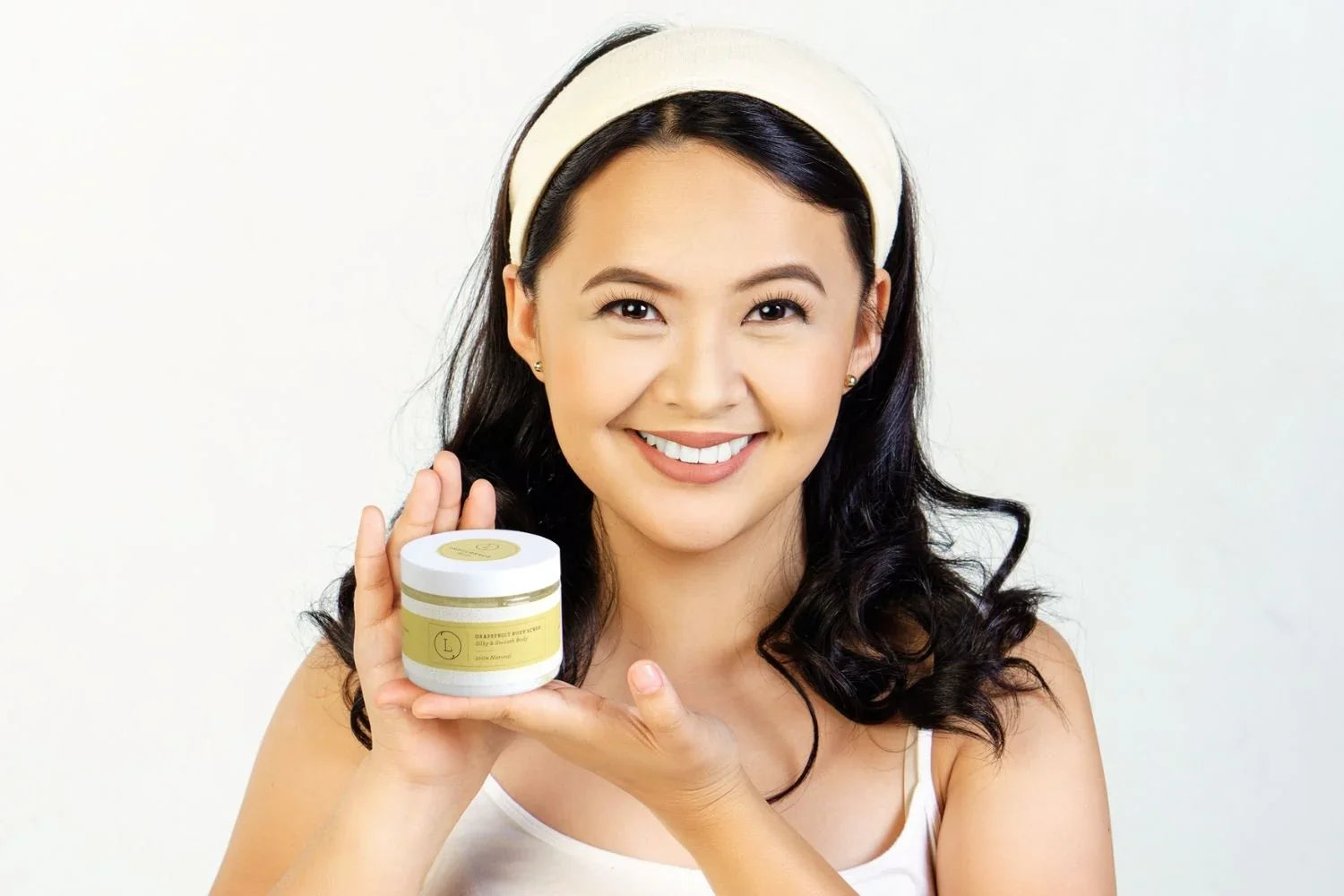 Smiling woman with headband holding grapefruit body scrub jar, natural skincare product
