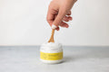 Hand holding wooden spoon scooping grapefruit body scrub from jar with yellow label on gray surface