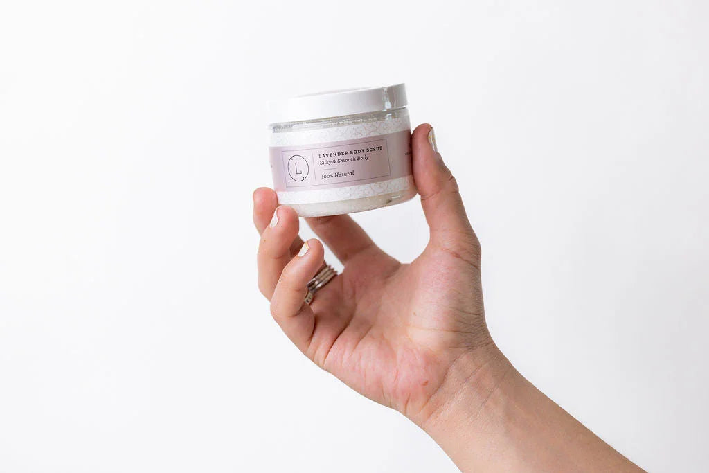 Hand holding a jar of lavender body scrub with a purple label against a white background