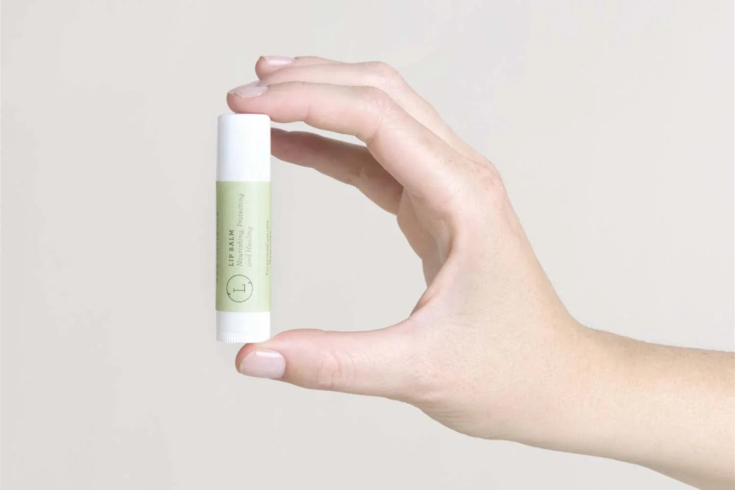 Hand holding a natural green label lip balm stick against a plain light background