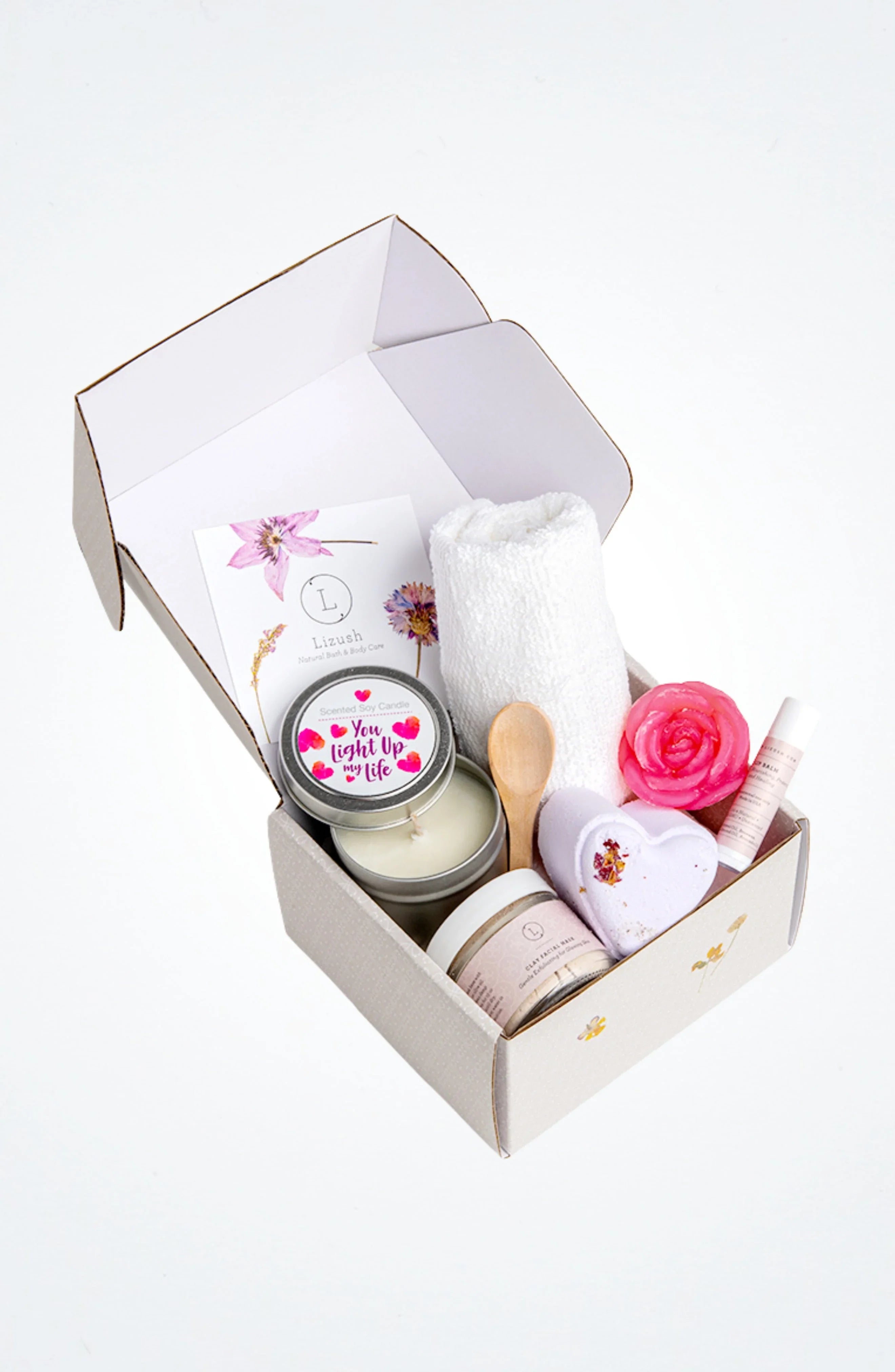 Open gift box with spa items including scented soy candle, rolled white towel, clay facial mask, heart-shaped bath bomb, lip balm, and wooden spoon