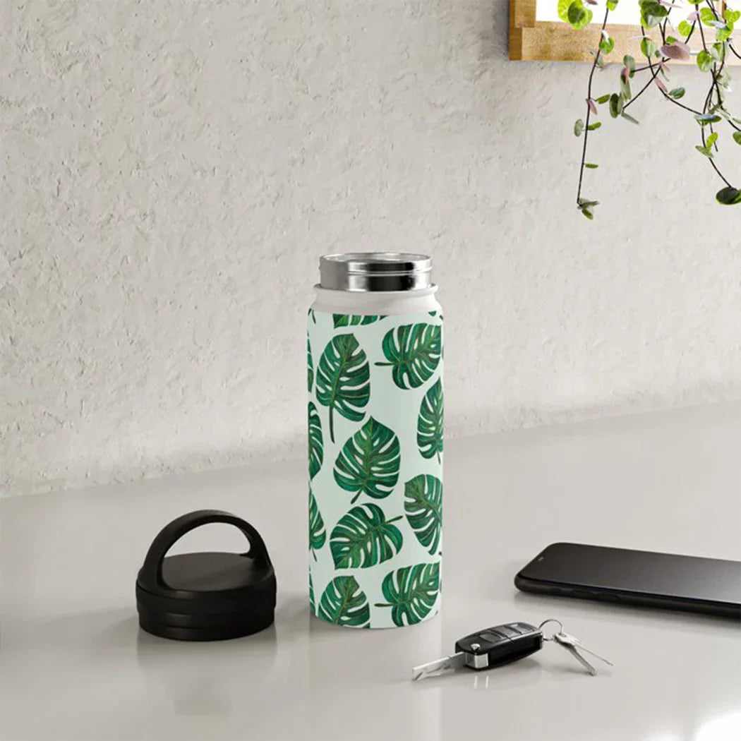 White insulated water bottle with green monstera leaf pattern, black cap, car keys, and smartphone on white table