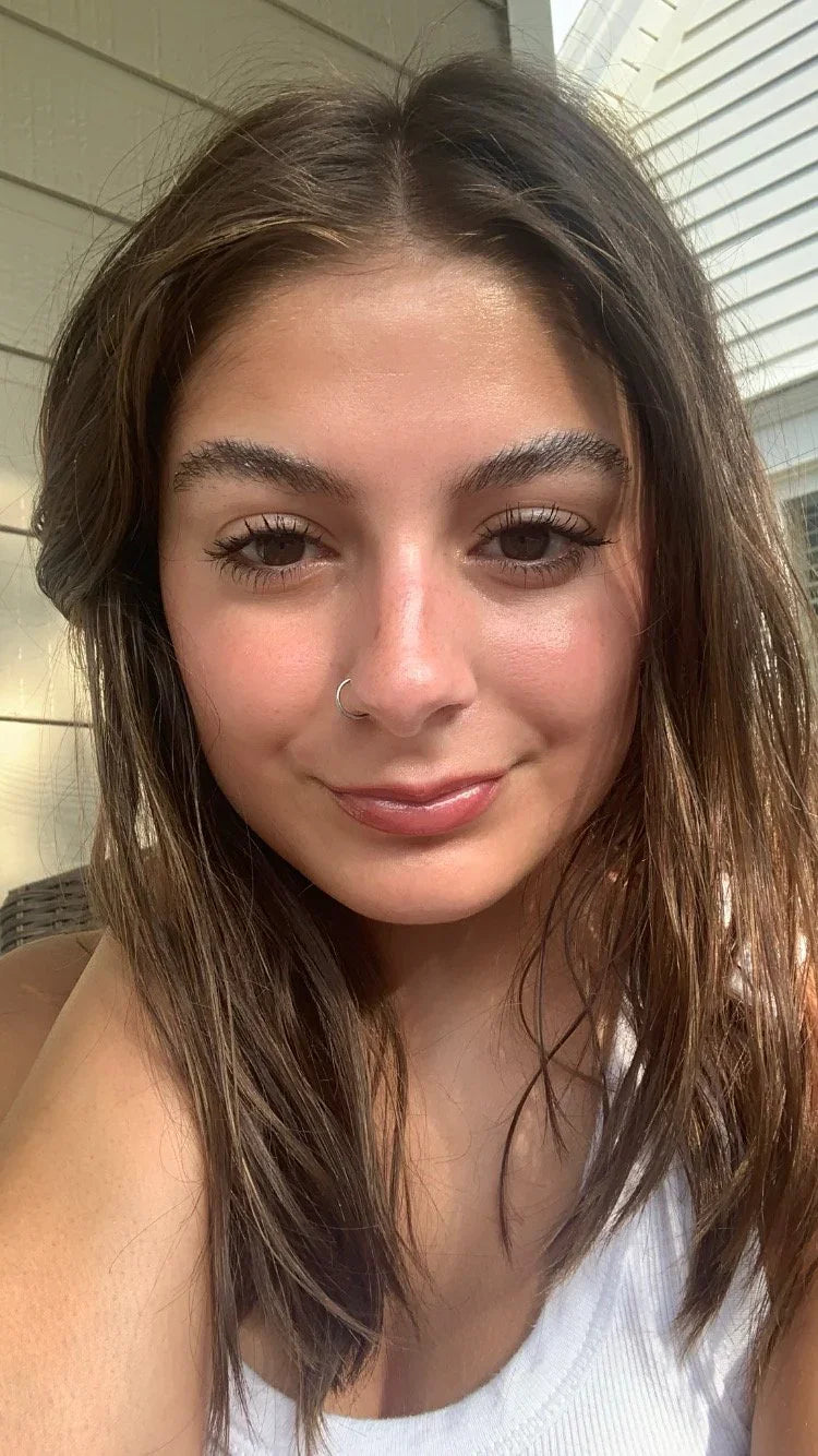 Young woman with brown hair, wearing a nose ring and white tank top, in natural daylight