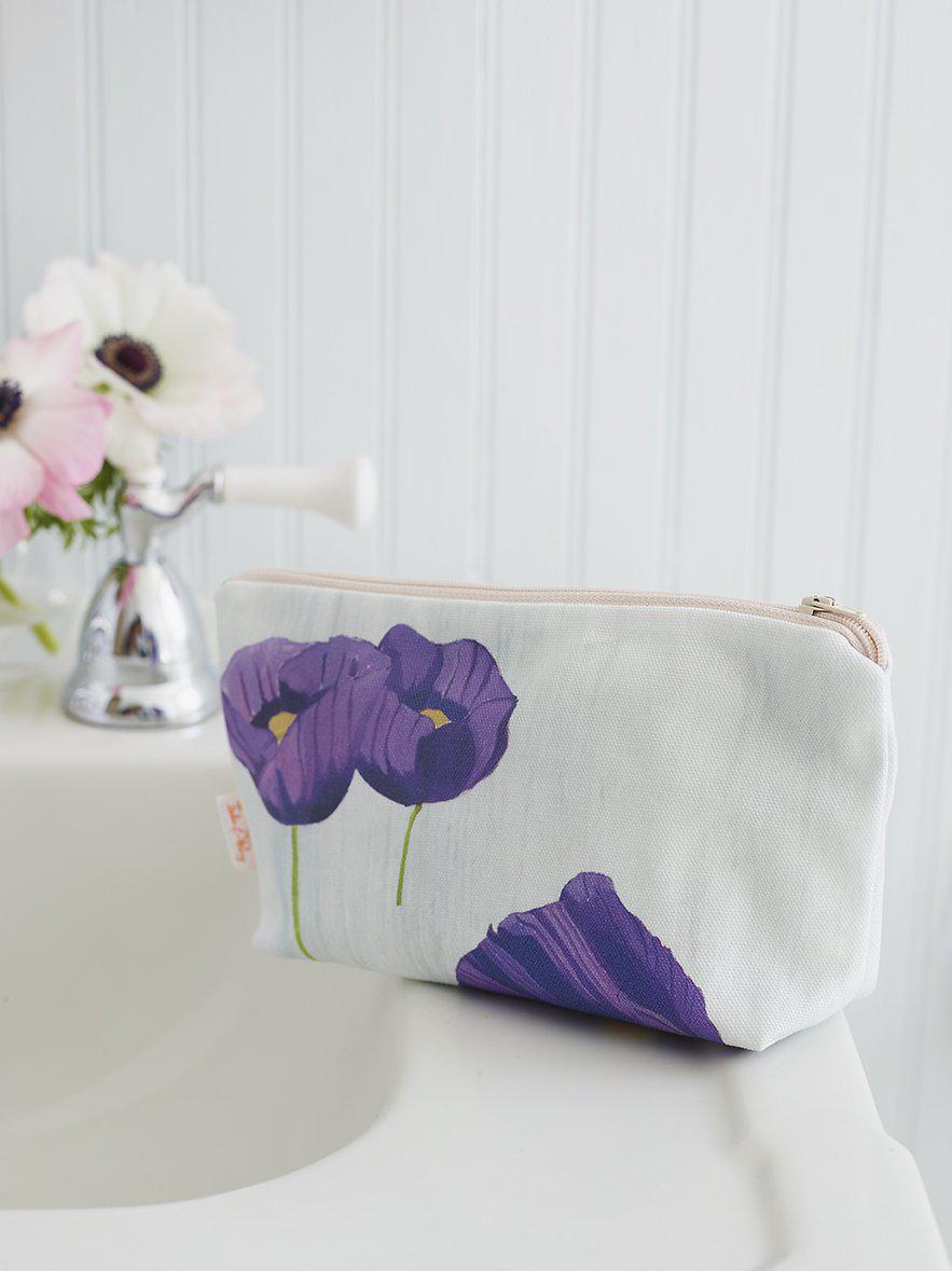White makeup bag with purple poppy flowers on white bathroom sink beside silver faucet and floral decor
