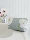 Grey fabric makeup bag with white poppy flowers on bathroom sink edge near chrome faucet