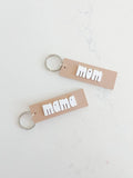 Tan keychains with bold white 'mom' and 'mama' text on a white marble surface, Qu!rk Boutique