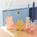 Modern pink sculptural vase with foliage, bold yellow vase, and blue candles on a wooden table