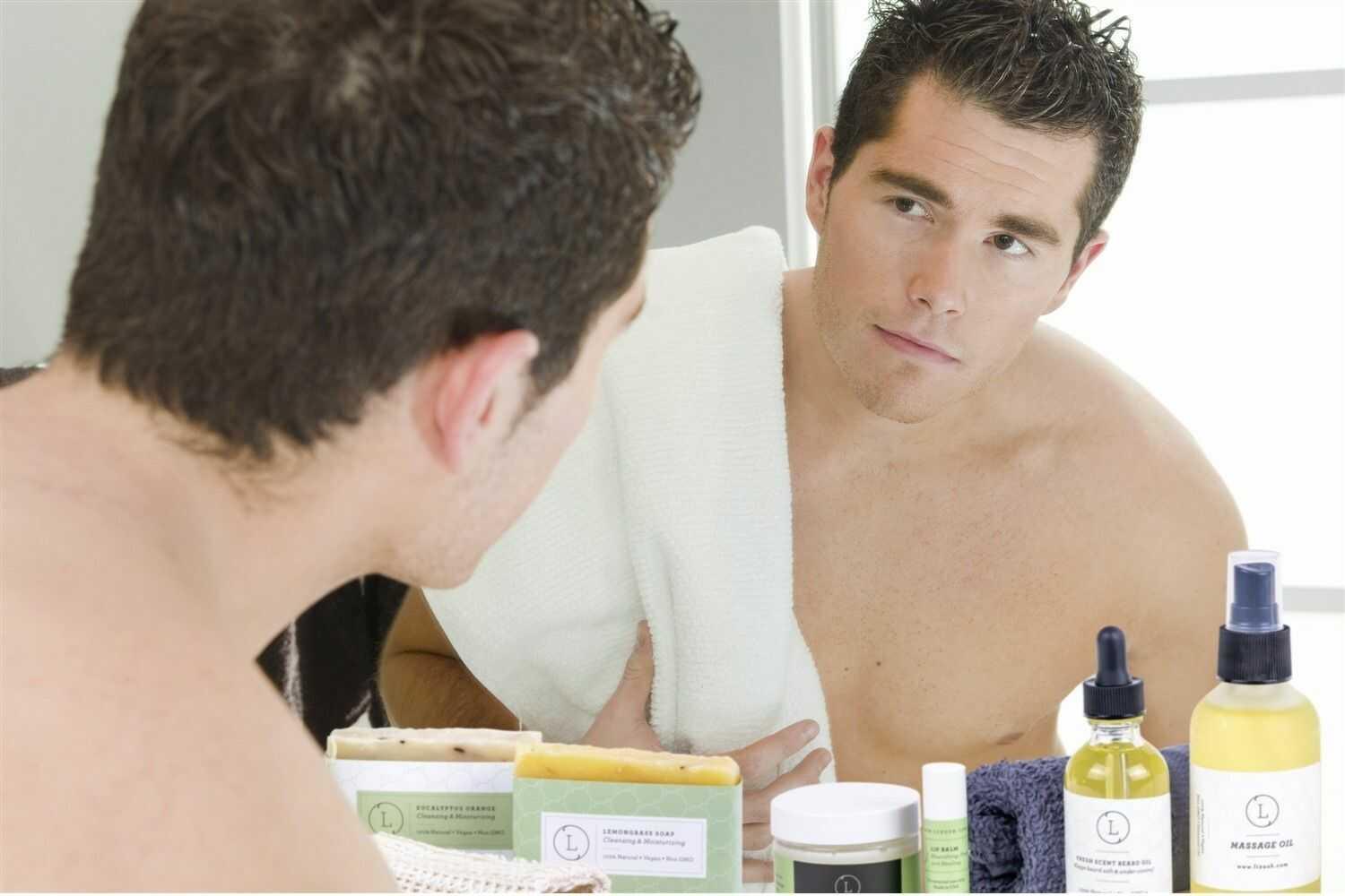 Man with towel on shoulder looking in mirror, surrounded by natural skincare products including soap, serum, balm, and massage oil