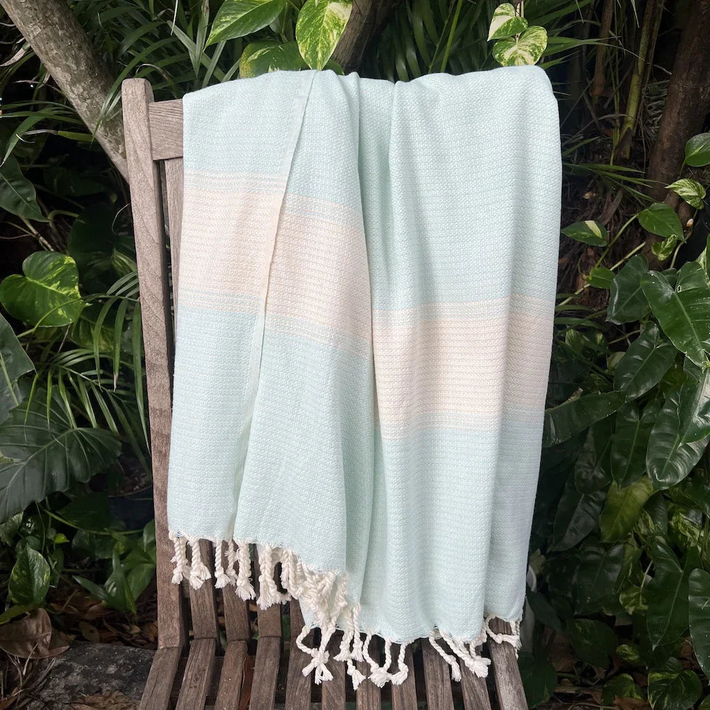 Mint Turkish towel with fringe draped on wooden chair, surrounded by lush green plants