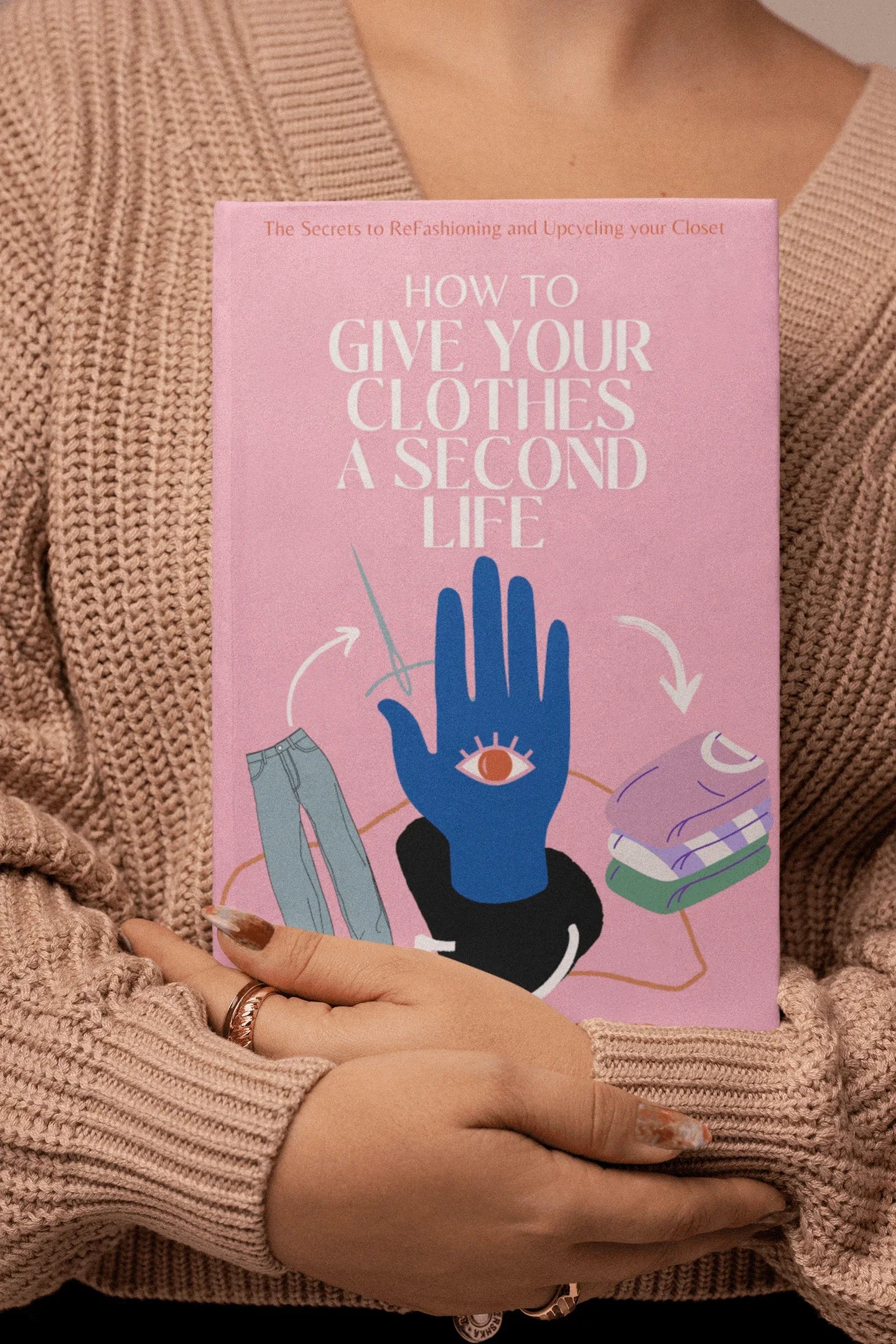 Person with long manicured nails holding a pink book titled 'How to Give Your Clothes a Second Life' in cozy beige sweater