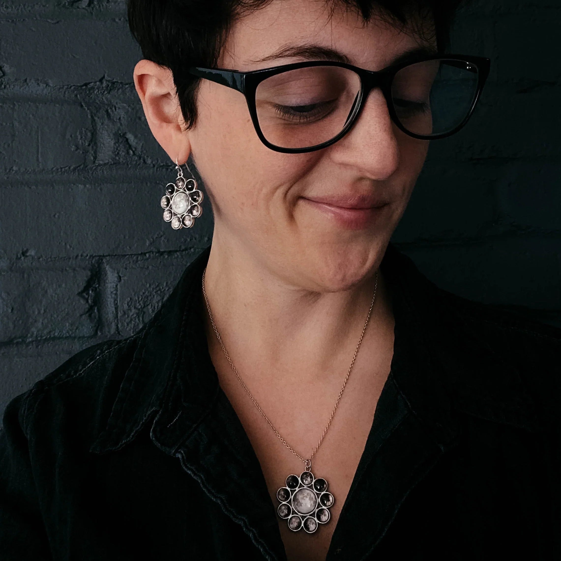 Person wearing black glasses and moon phase pendant necklace and matching earrings against dark brick wall