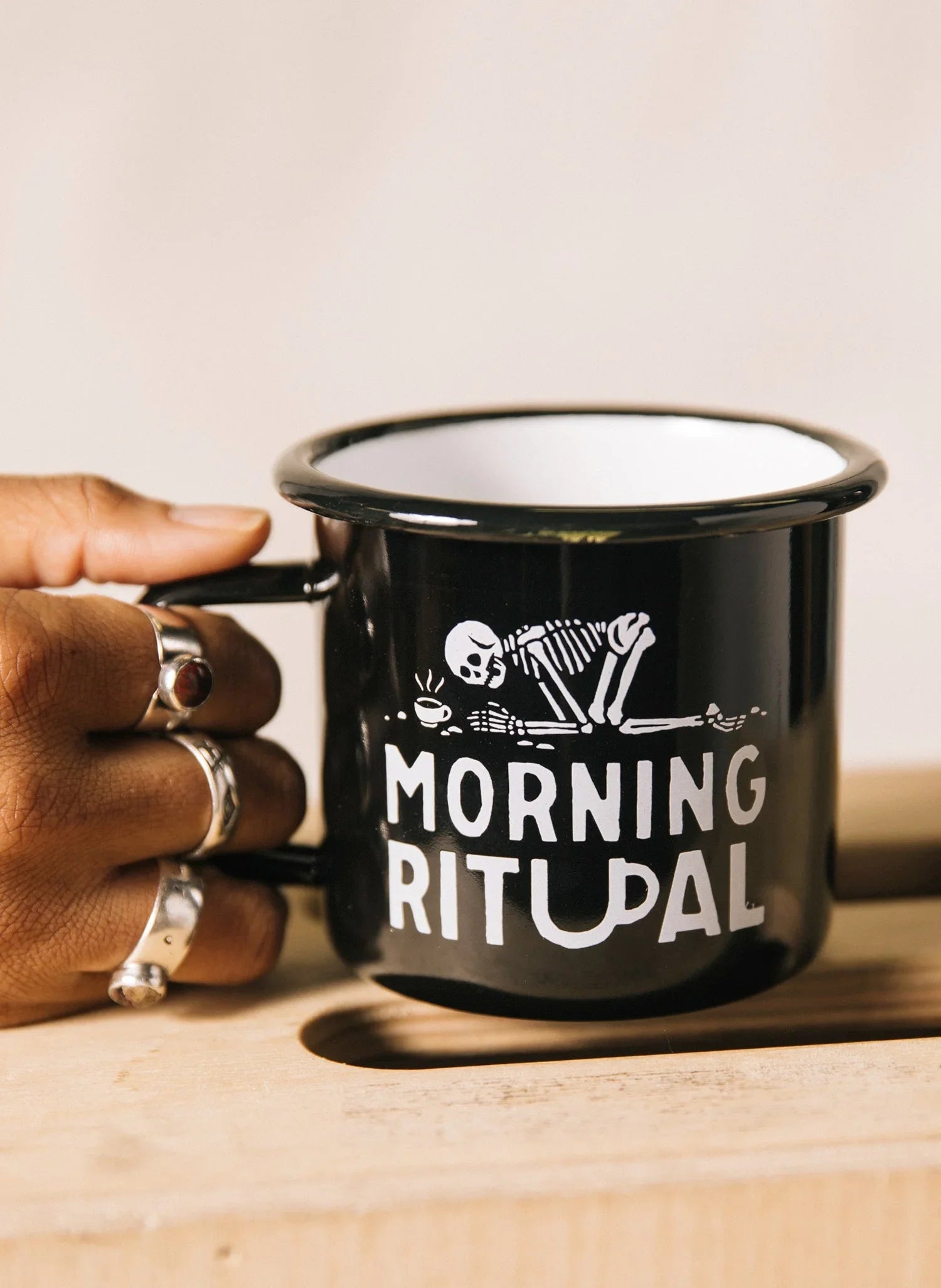 Black enamel mug with white 'MORNING RITUAL' text and skeleton graphic, held by a hand with rings, from Qu!rk Boutique.