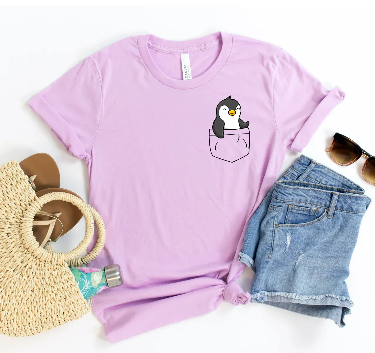 Lavender t-shirt with a cute penguin peeking from a printed pocket, paired with denim shorts, sandals, sunglasses, and straw bag