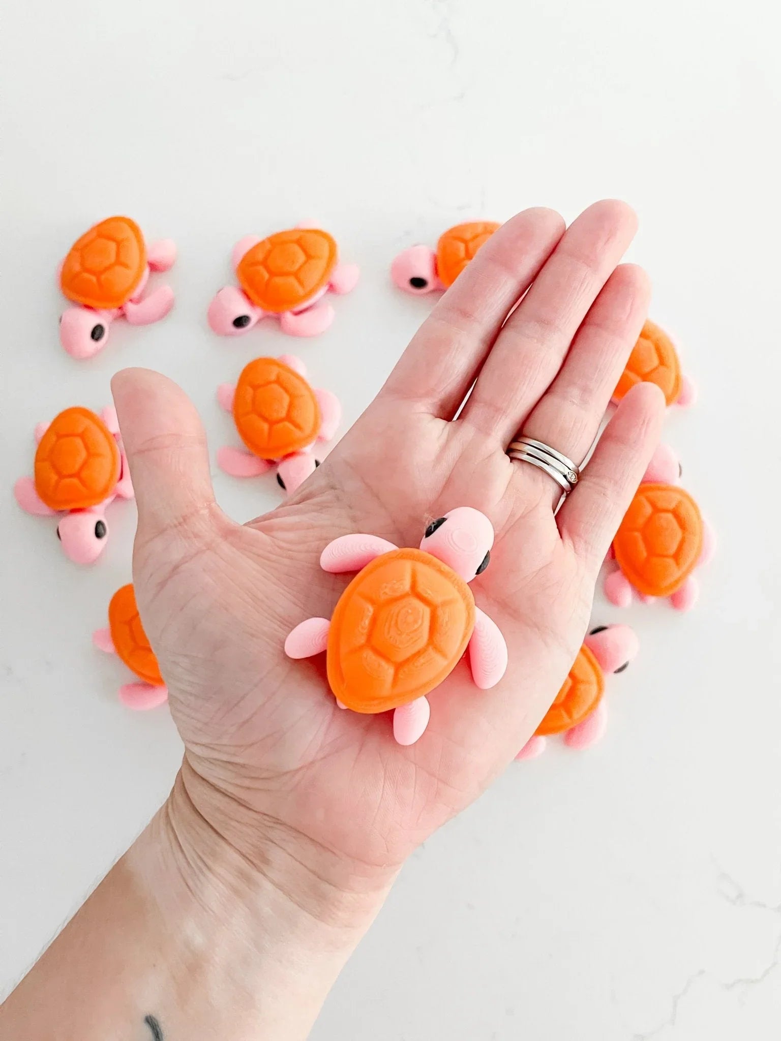 Hand holding a small orange and pink turtle toy with more toy turtles on a white surface