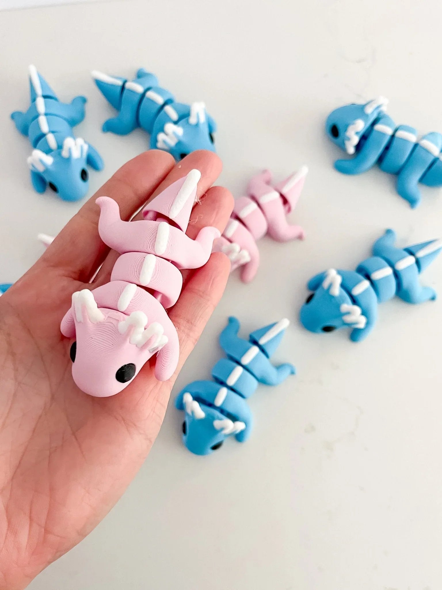 Hand holding a pink axolotl baby critter toy among blue and pink axolotl figurines on white background.