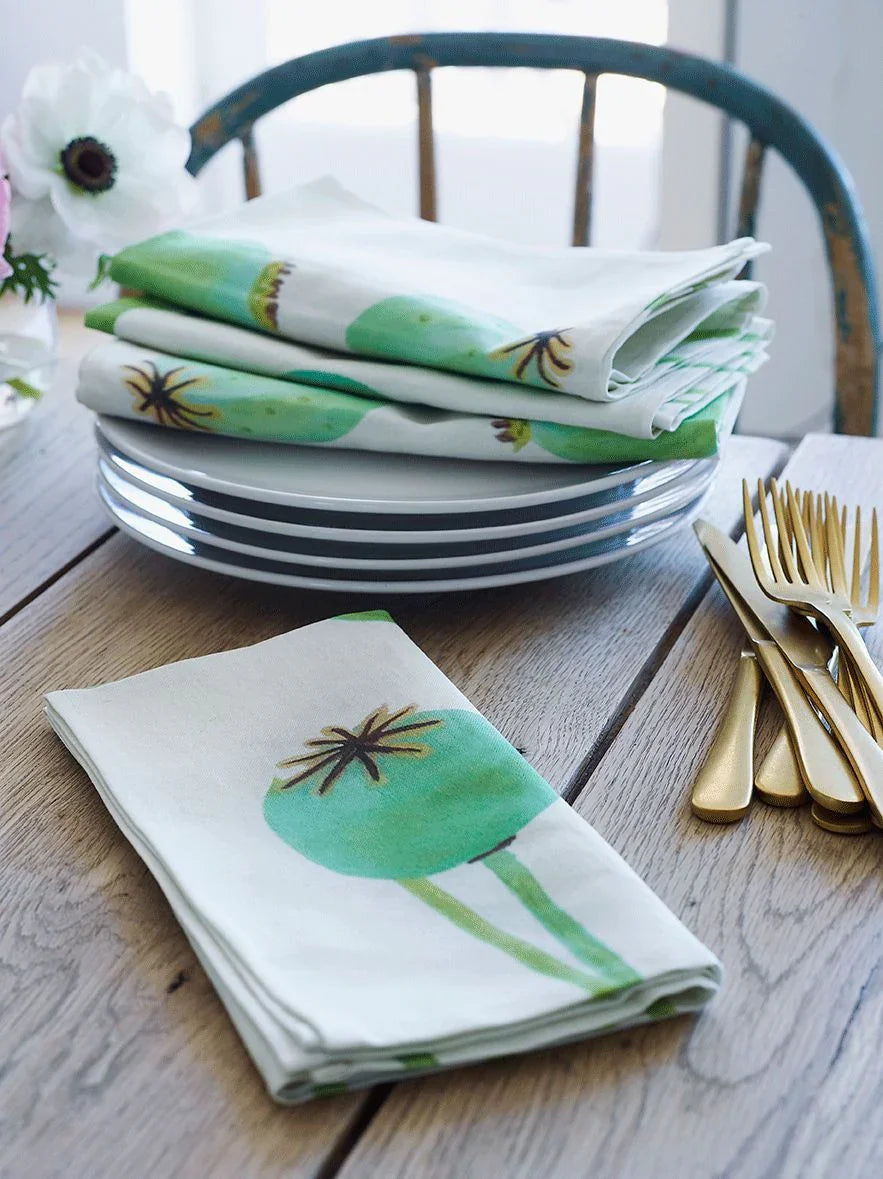 Folded cotton napkins with green poppy pod print on ecru linen, stacked on white plates with gold cutlery