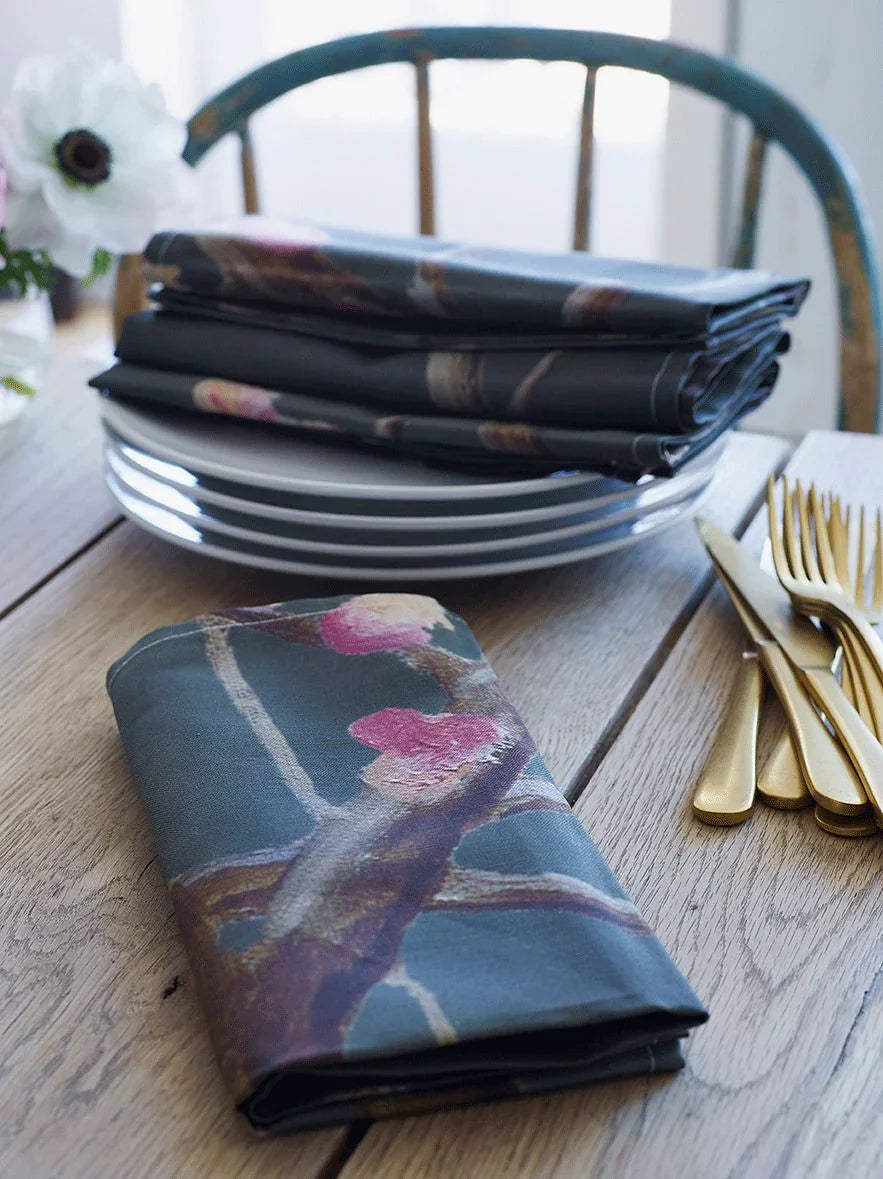 Folded dark grey napkins with pink flower tree design on wooden table with stacked white plates and gold cutlery