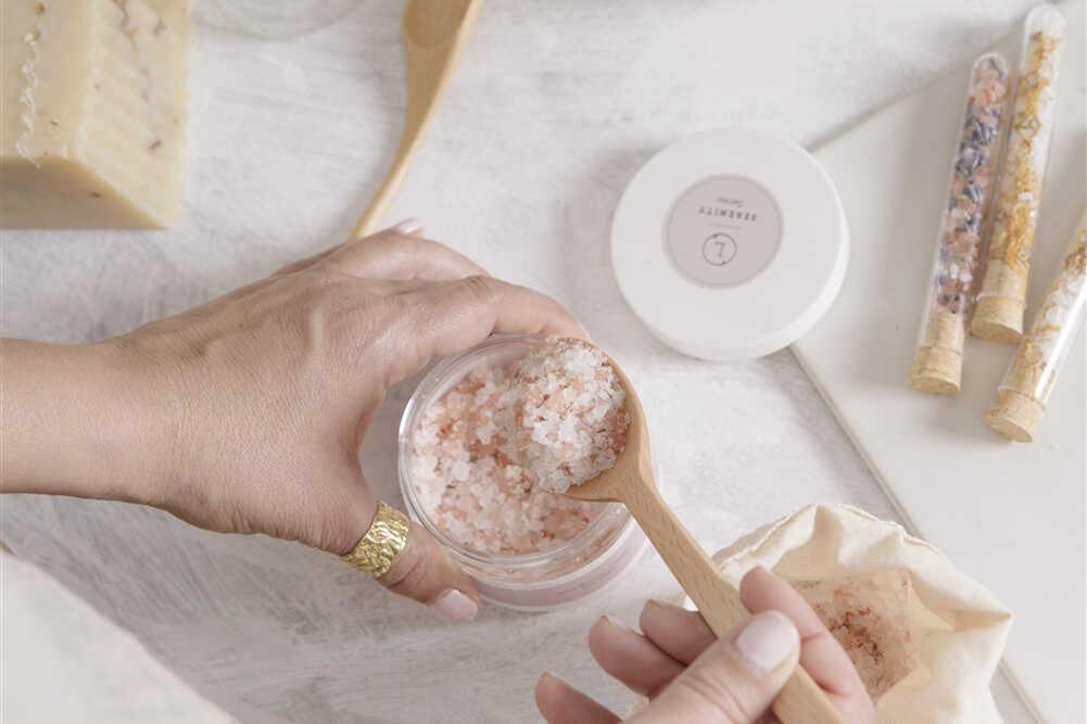 Hands scooping pink natural bath salt from a jar with wooden spoon on white surface, spa setup