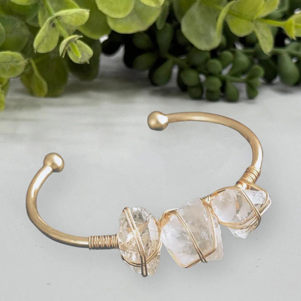 Gold wire-wrapped crystal cuff bracelet on marble surface, greenery in background, Qu!rk Boutique