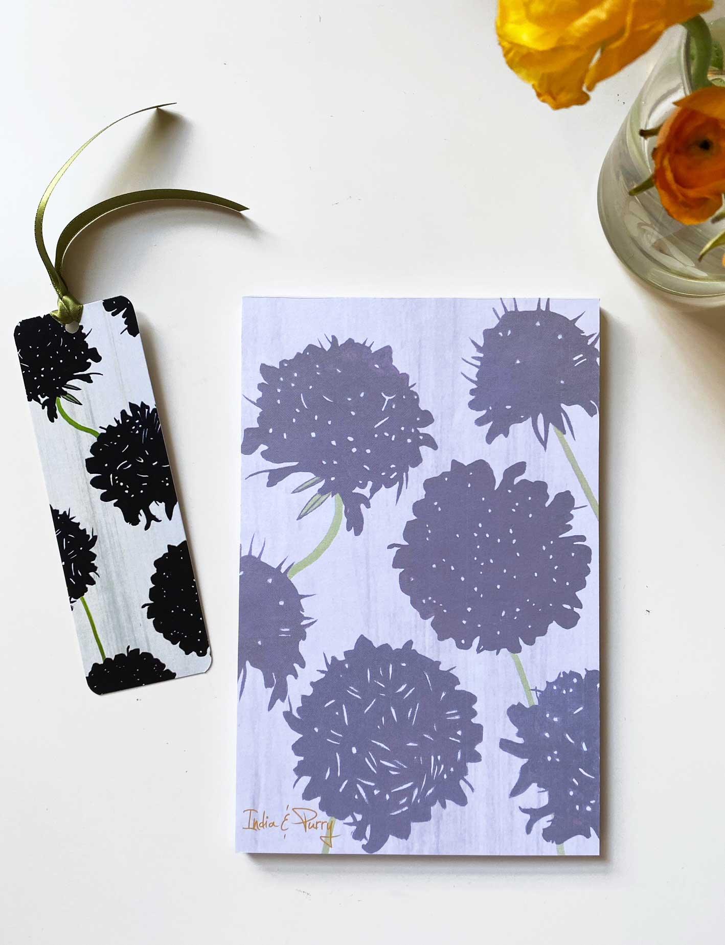 Notepad and bookmark with black scabiosa flower print on white surface, yellow flowers in vase