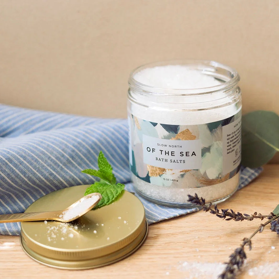 Jar of Slow North Of The Sea bath salts with gold lid, spoon with salts, green mint leaves, and dried lavender on wood