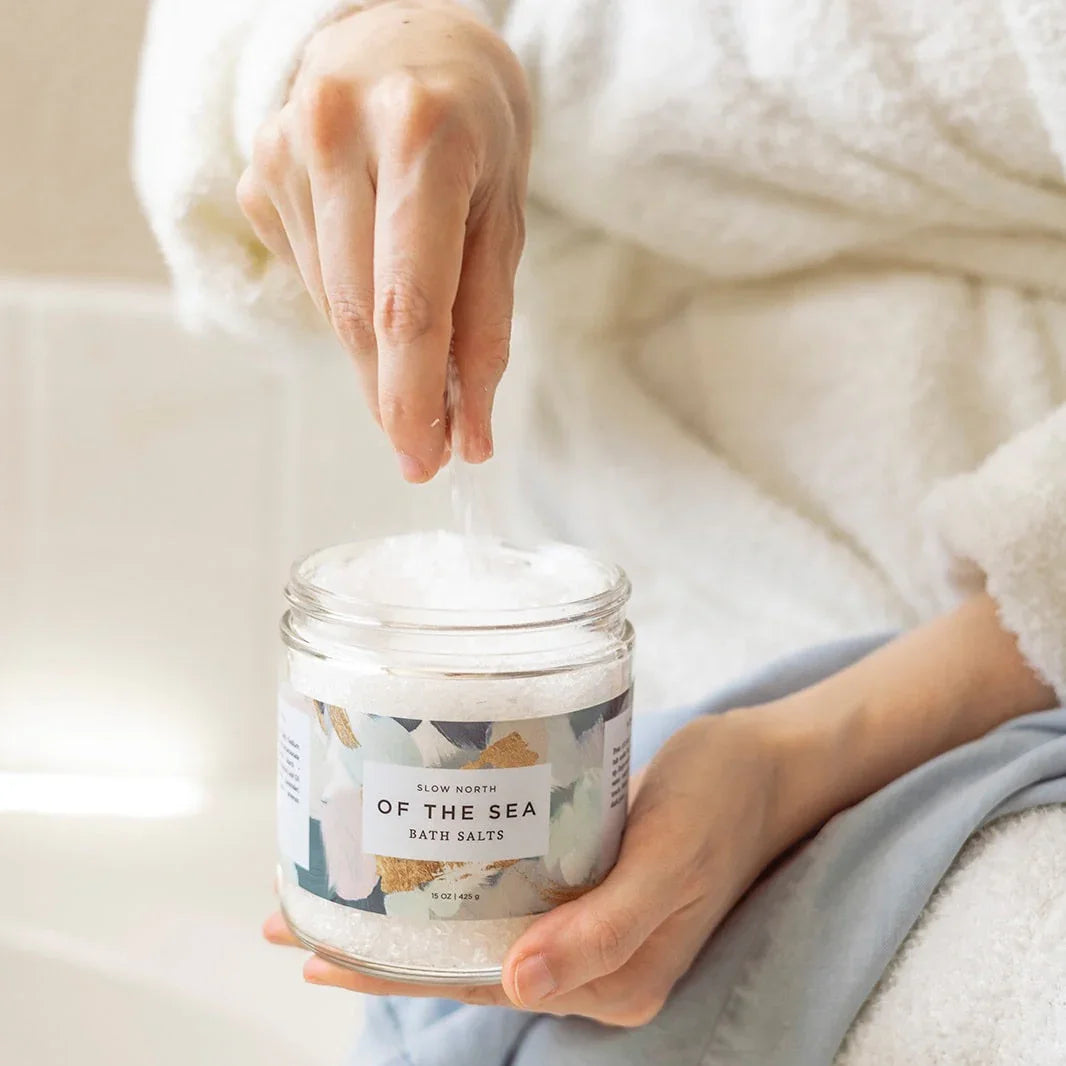 Person wearing a white robe sprinkling Slow North Of The Sea bath salts from a glass jar