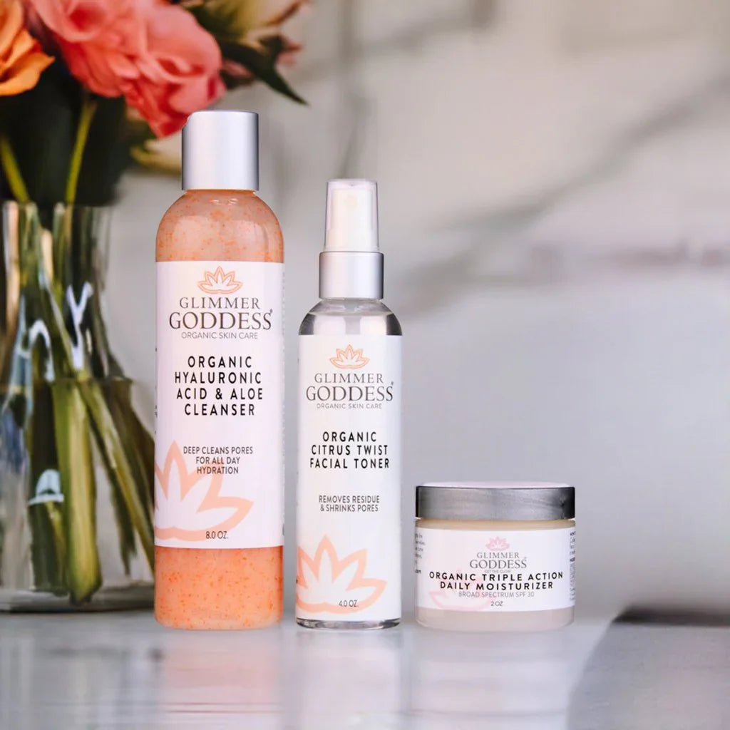 Glimmer Goddess organic 3-step anti-aging skin care kit with cleanser, toner, and moisturizer on a counter, flowers blurred in background