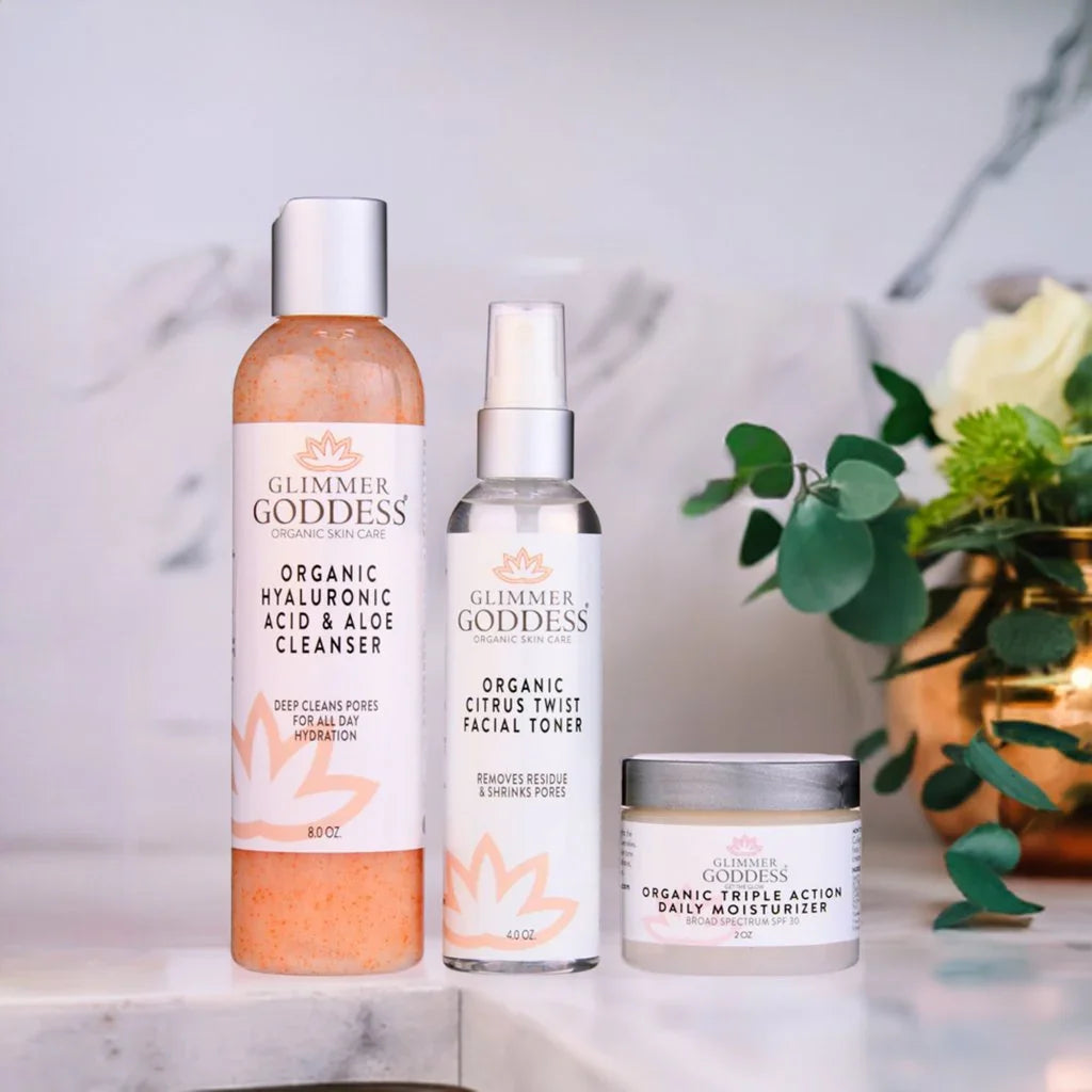 Glimmer Goddess organic 3-step anti-aging skin care kit with cleanser, toner, and moisturizer on marble counter next to flowers at Qu!rk Boutique.
