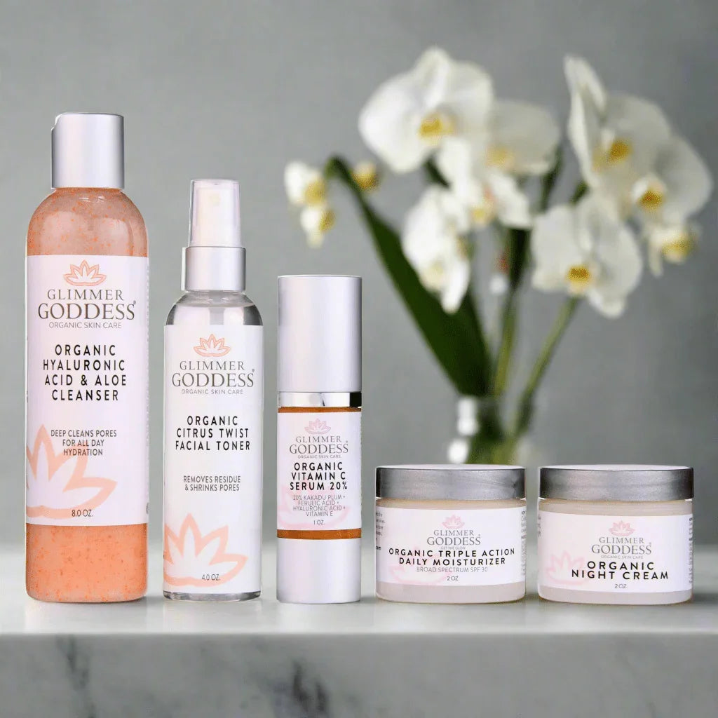 Glimmer Goddess organic skincare set including cleanser, toner, vitamin C serum, moisturizer, and night cream on marble surface