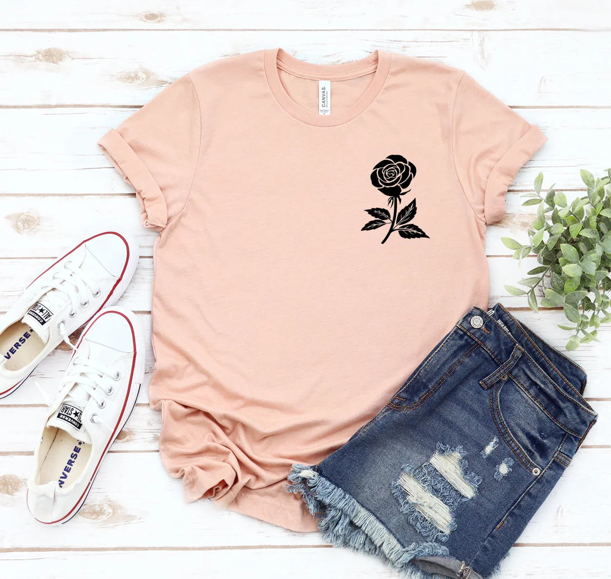 Pink rolled-sleeve t-shirt with black rose print, distressed denim shorts, white Converse sneakers on white wood