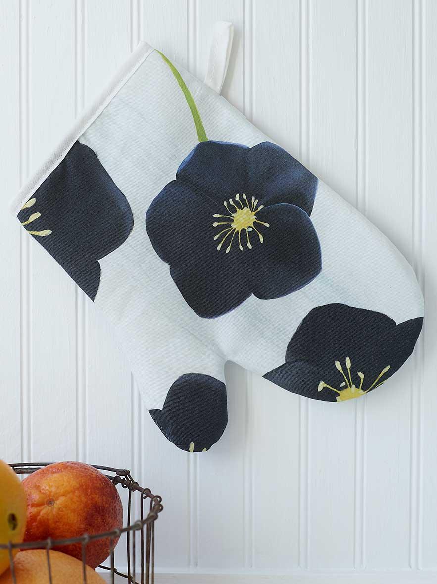 Black flower-patterned oven mitt hanging on white paneled wall above wire basket with oranges
