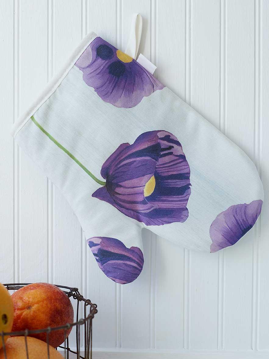 White oven mitt with purple poppy flower design hanging on white paneled wall next to fruit basket