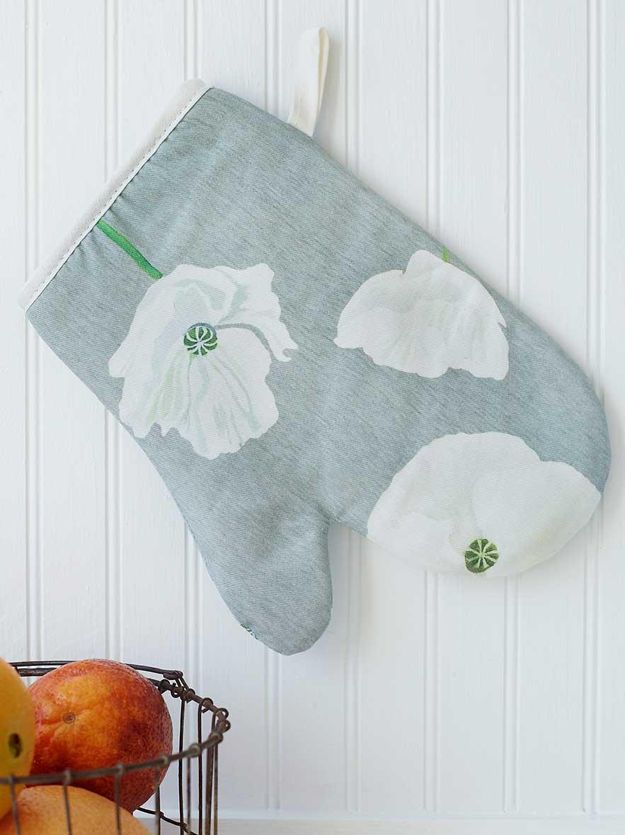 Gray oven mitt with white poppy flower print hanging on white beadboard wall with fruit basket nearby