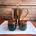Hammered copper pitcher with lid and two artisan cups on a white cloth at Qu!rk Boutique