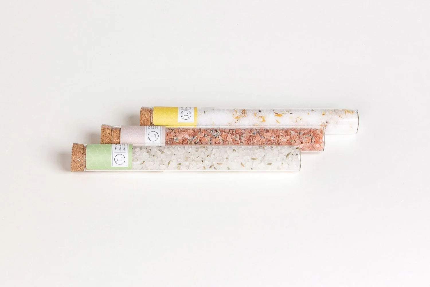 Three glass tubes with cork stoppers filled with bath salts of different colors and herbs on a white background