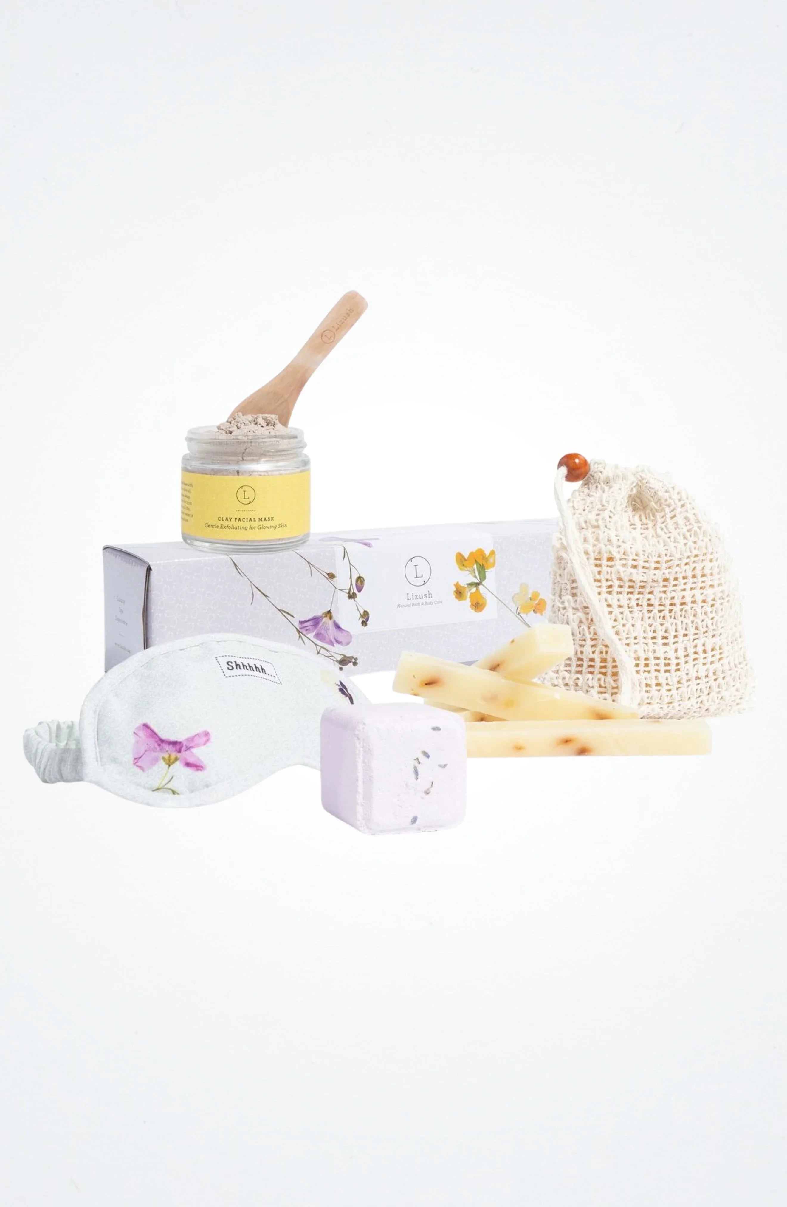 Pampering kit with clay facial mask, floral eye mask, bath bomb, soap bars, and mesh bag on white background