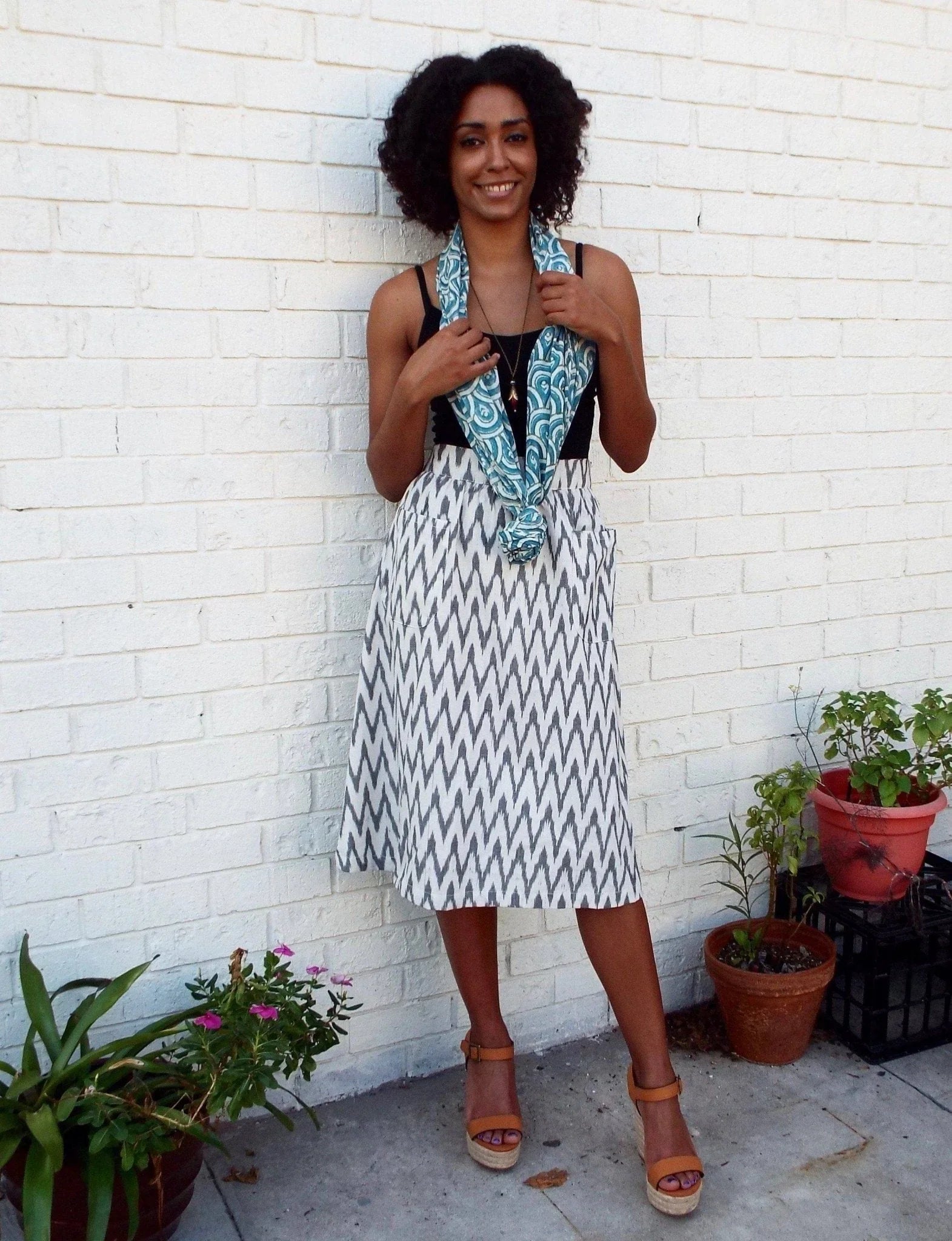 Smiling woman wearing teal peacock scarf, patterned skirt, and sandals by white brick wall.