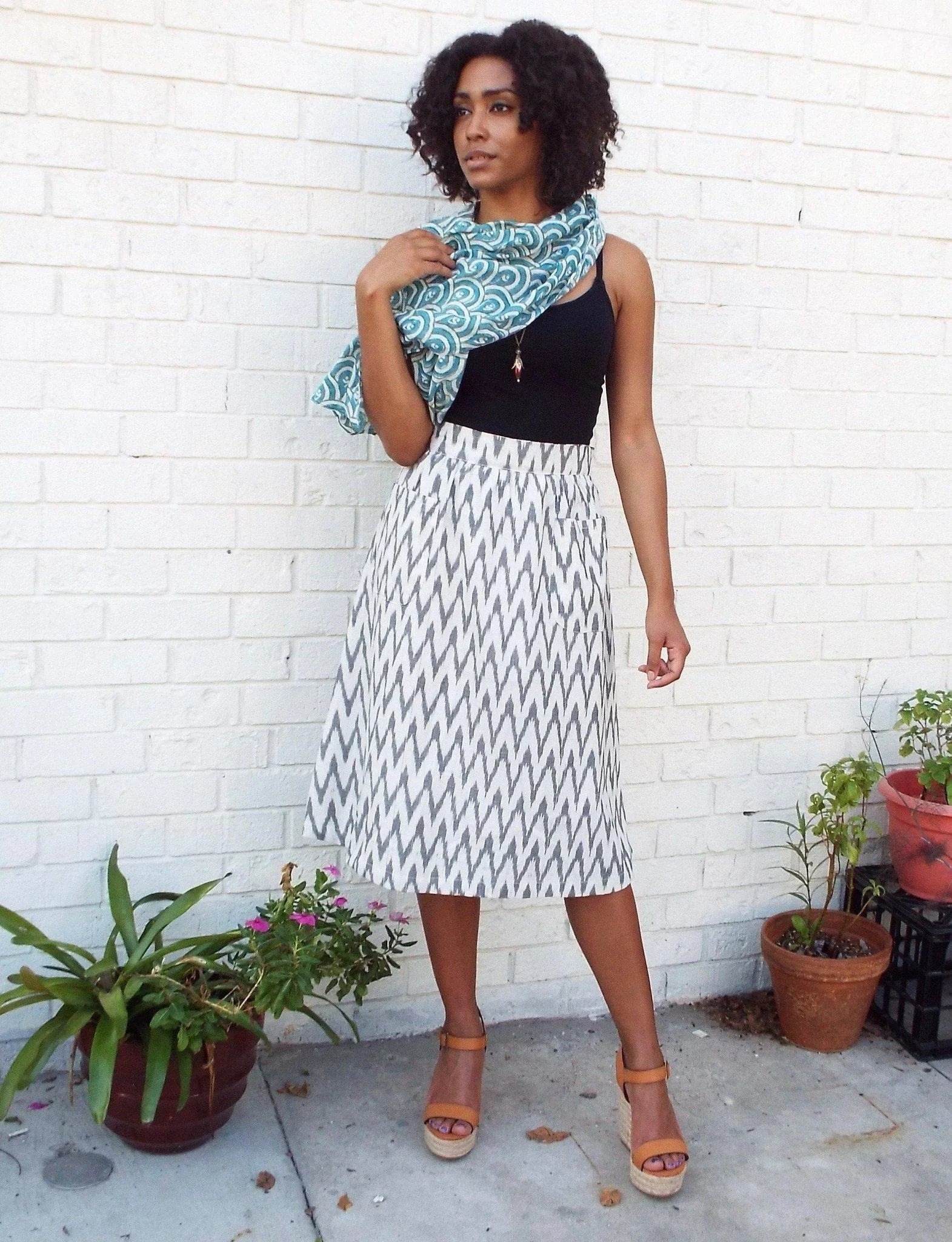 Woman modeling teal peacock-pattern scarf, zigzag skirt, and sandals by white brick wall