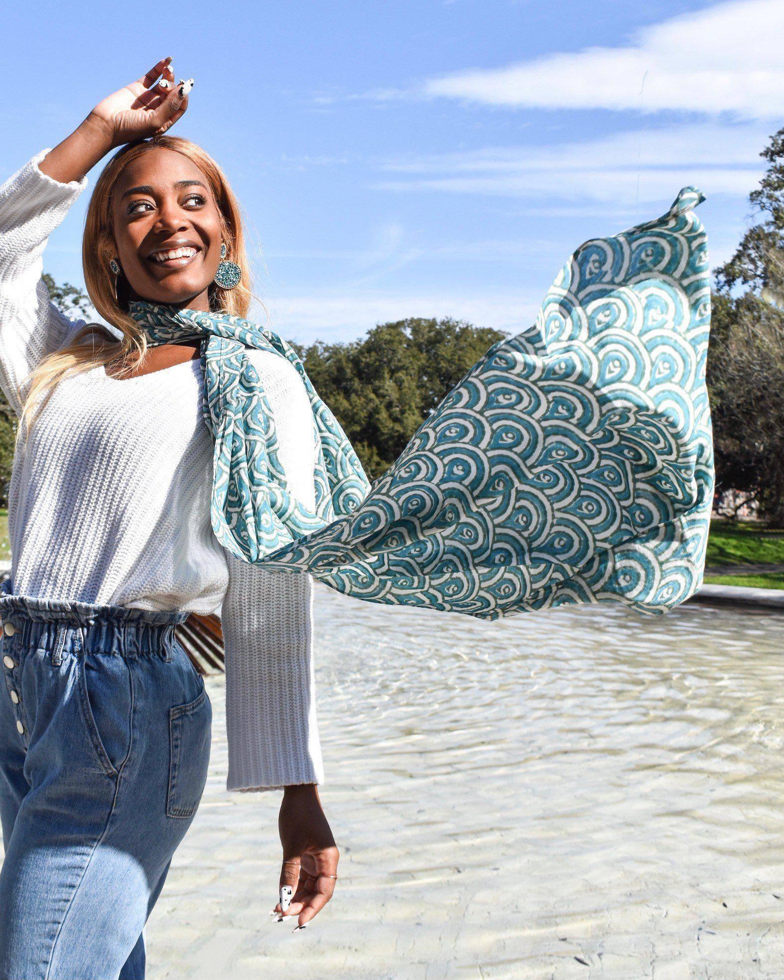 Smiling woman outdoors in white sweater and jeans, wearing a teal peacock-print scarf from Qu!rk Boutique.