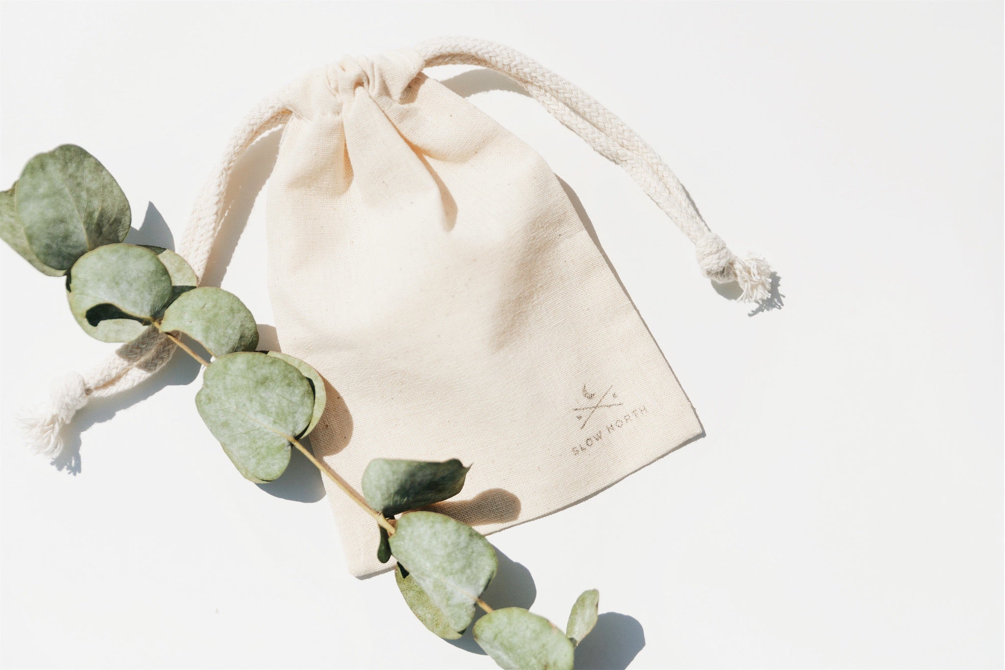 Natural cotton drawstring pouch with Slow North logo beside eucalyptus branch on white background