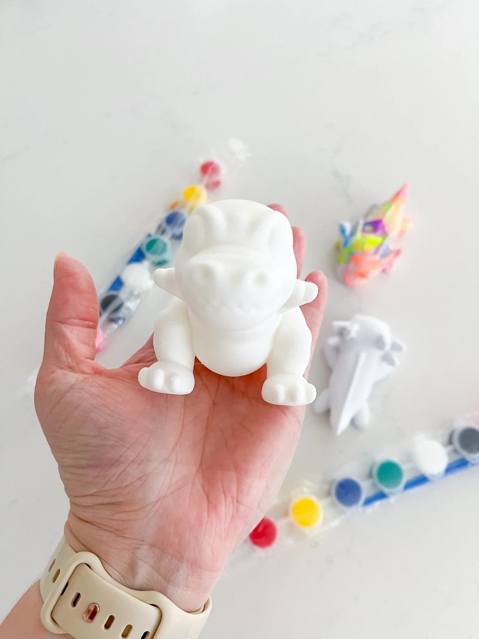 Hand holding a white dragon figurine from a paint-your-own kit with paint pots and brush nearby