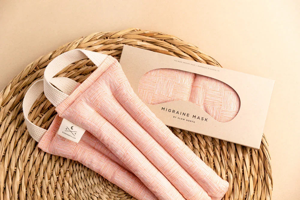 Pink woven migraine masks with natural strap handles on a wicker tray, shown with matching packaged eye mask