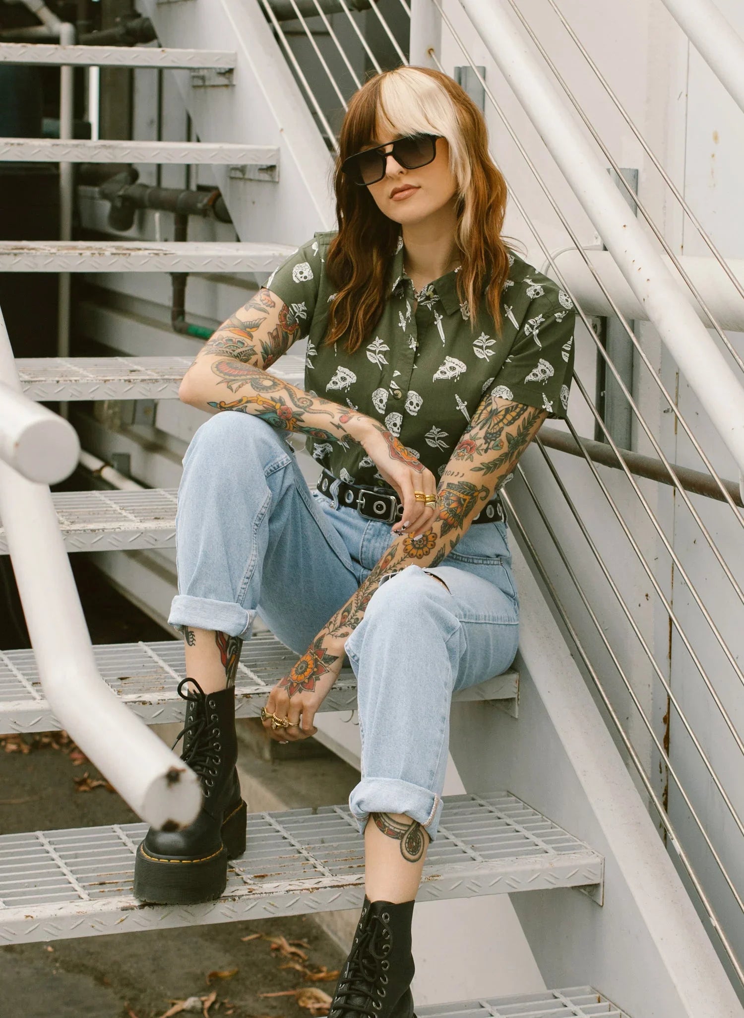 Tattooed woman in sunglasses, skull print button-up shirt, and cuffed jeans on outdoor stairs